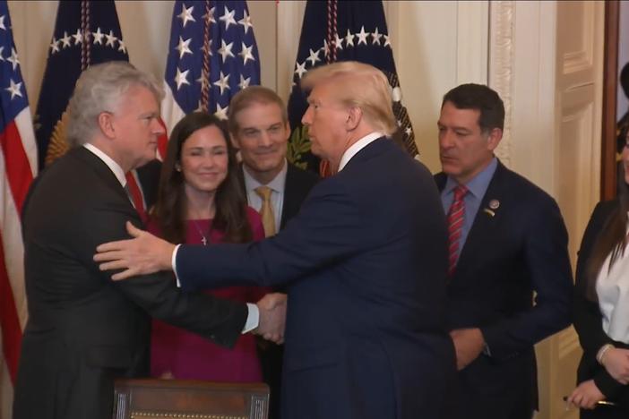 President Donald Trump is shown shaking the hand of Rep. Mike Collins while the family of Laken Riley and others look on, in a White House setting surrounded by American flags.