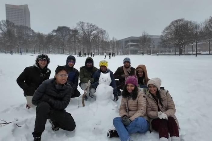 Nine students in winter clothing are shown outside in a snowy field seated on the ground near a snowman.