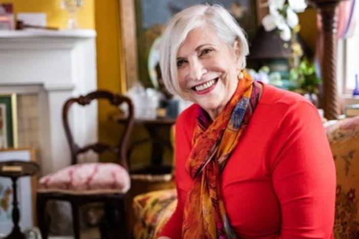 Nathalie Dupree is shown smiling and seated inside a floral and colorful domestic setting.