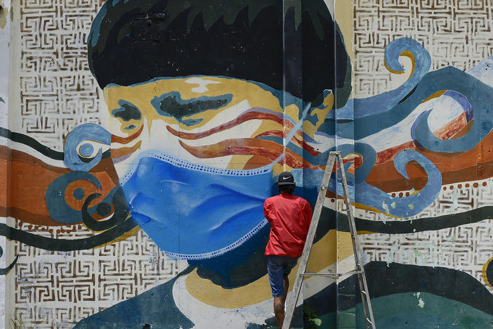 A street artist spray paints a protective face mask over an old mural featuring a Venezuelan Indigenous man, in Caracas, Venezuela, on Saturday. Globally, new daily cases hit an all time high, the World Health Organization reports.