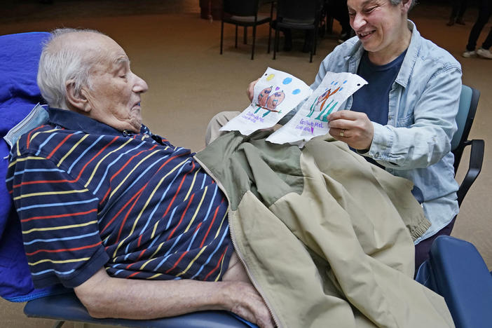 Melvin Goldstein, 90, glances at pictures of birds, left, and a fish, his 13-year-old granddaughter drew for him as a gift as his daughter Barbara Goldstein shares them with his during a family visit inside the Hebrew Home at Riverdale on March 28 in New York. On Friday, the government eased many remaining pandemic restrictions.